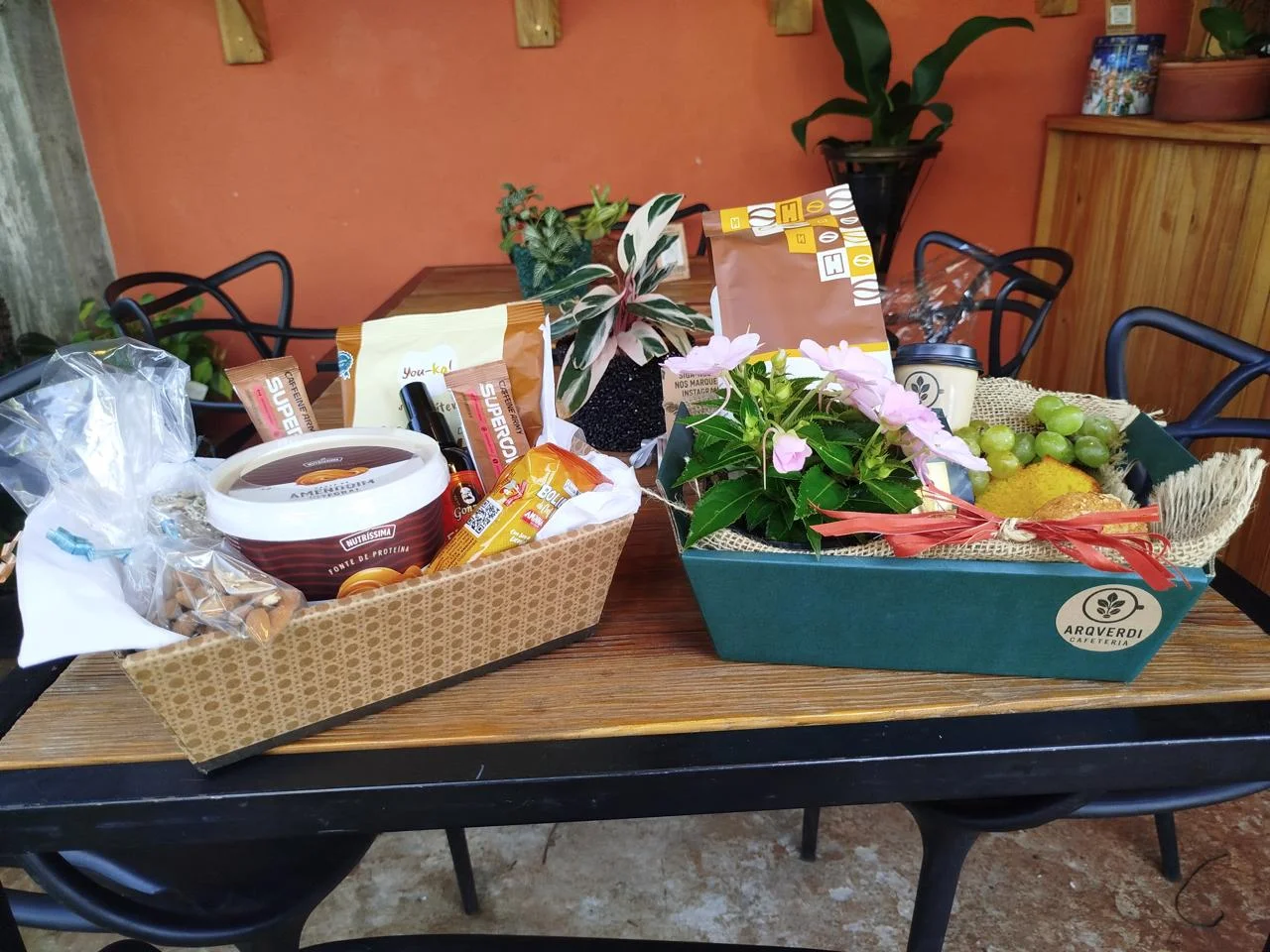 Cestas ilustrativas da ArqVerdi Cafeteria com produtos de café da manhã e plantas sobre uma mesa de madeira.